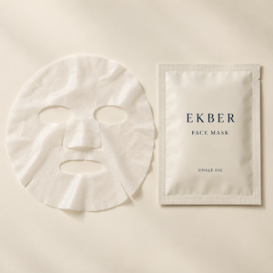 Thin white single-use facial sheet mask beside an ivory Ekber sachet